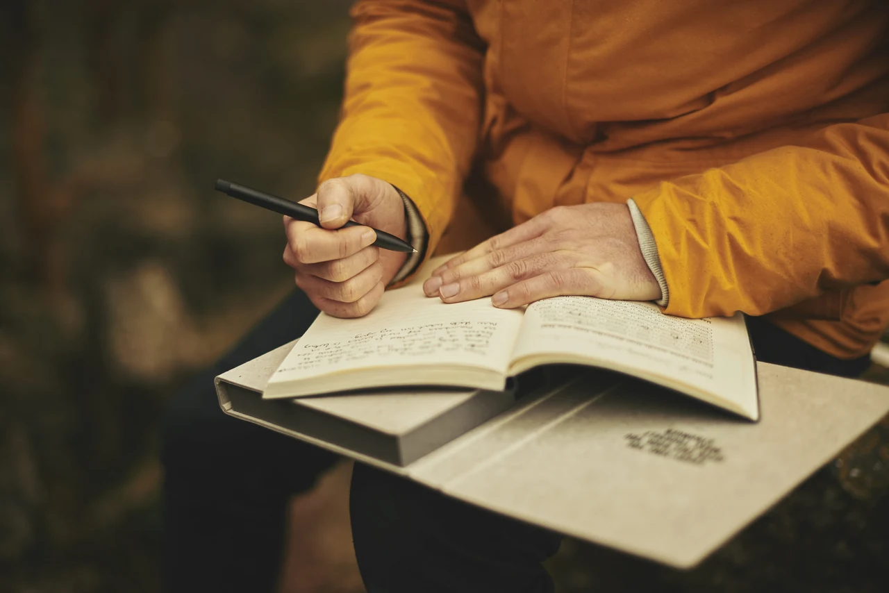 Nahaufnahme: Hände schreiben in ein Notizbuch auf einer Klemmplatte, im Hintergrund unscharfe Natur