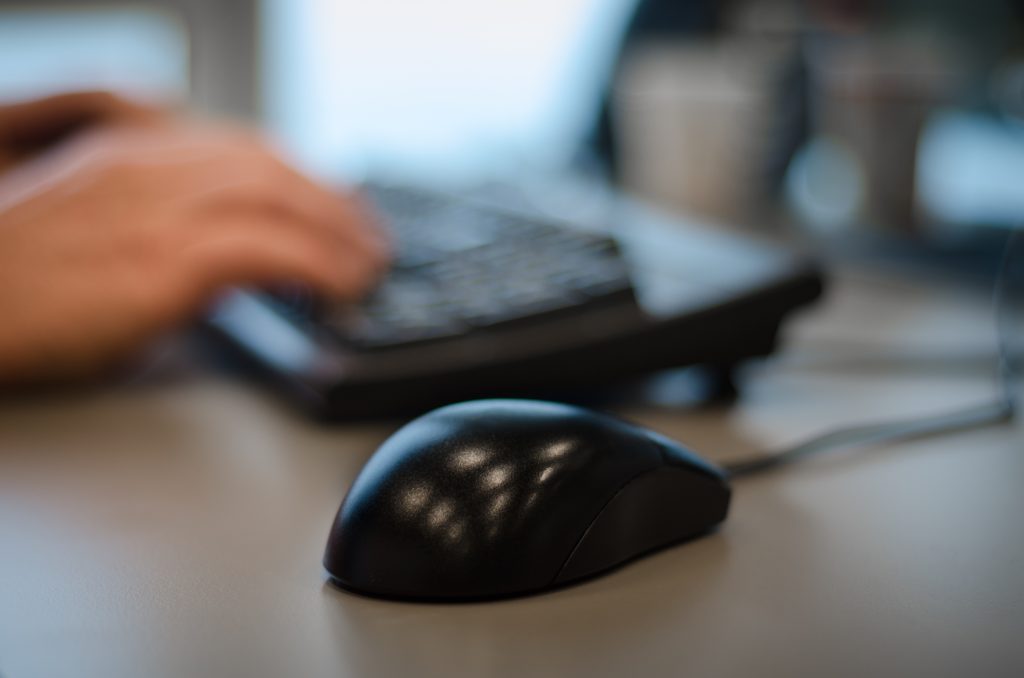 Hands typing on a keyboard, mouse in the foreground, blurred office in the background
